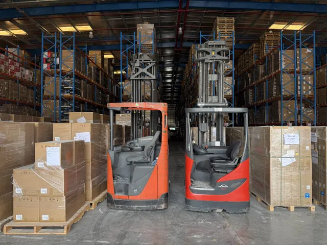 Two red electric reach trucks parked in a warehouse aisle beside stacked cartons